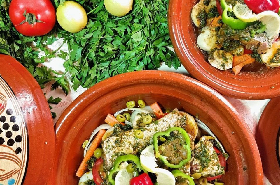 Overhead photo of tagine filled with fish and vegetables. Parsley,, lemon and tomato are on the table next to the tagines.