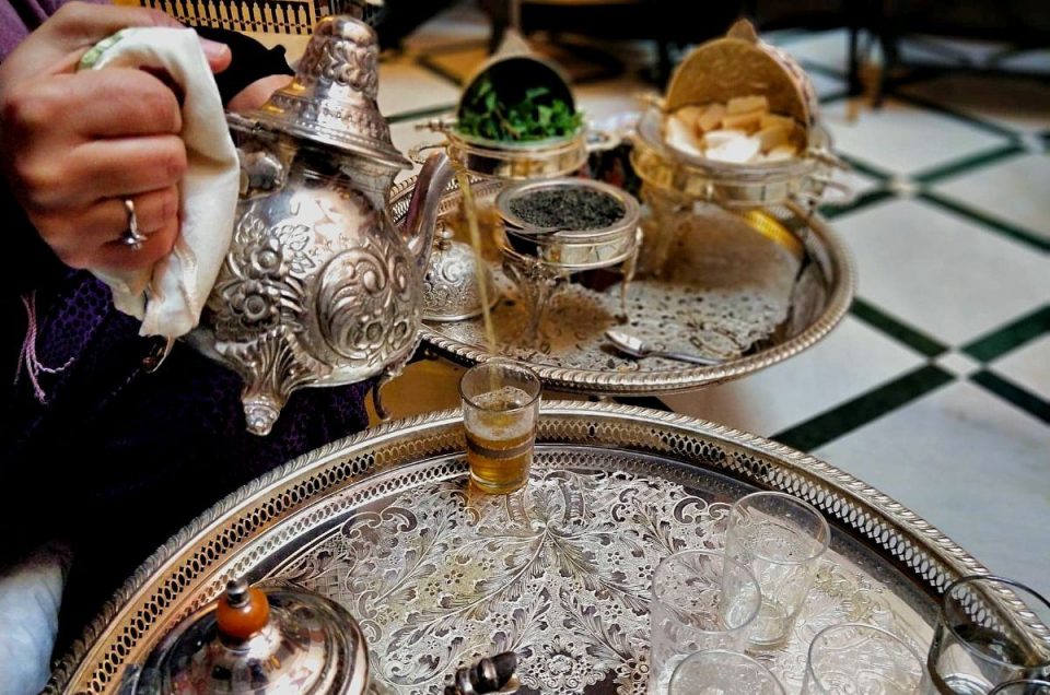 Closeup image of tea pouring from the long spout of an engraved metal Moroccan tea pot. The tea glass sits on an engraved metal tray with another tray and engraved tea, sugar and mint dishes in the background.