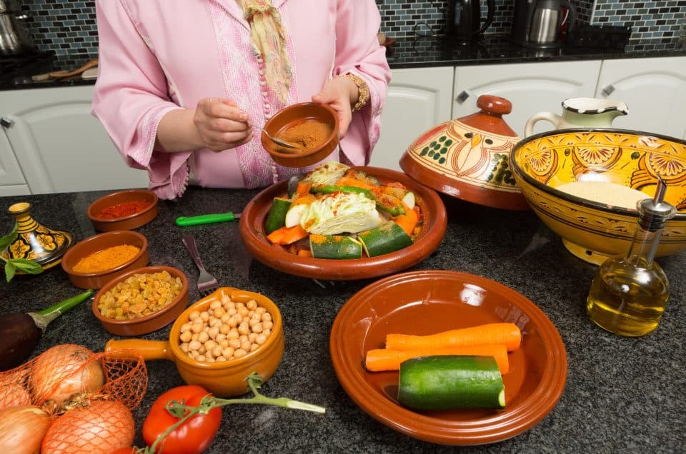 The torso of a woman is visible as she spoons spices into a tagine filled with vegetables.